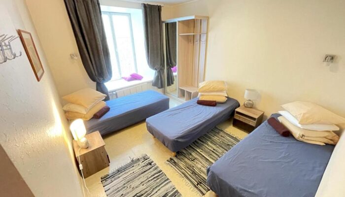 Room with 3 parallel beds. There is a wardrobe and a large window with curtains, bedside tables and a small lamp