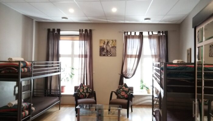 Big room with classic furniture. There are bunk beds on the left and right, and armchairs in the middle. The background wall has two large windows with curtains